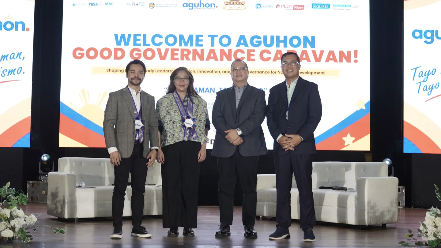 Atty. Third Bagro stands with three fellow panelists on stage, in front of a screen reading “Welcome to Aguhon Good Governance Caravan”